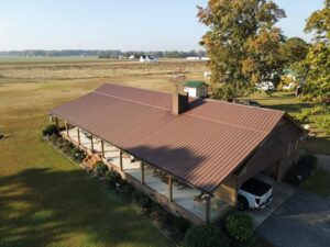 Aerial view of a large, single-story house with a brown halo roofing metal roof and a long wrap-around porch. The house is surrounded by a lawn with scattered trees and shrubs. Farmland and other small structures are visible in the background.