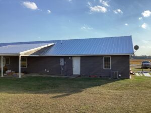 A single-story house with a dark exterior and a metal roof is shown, showcasing impeccable roofing work. The house has a covered patio area on the left side. Various construction materials are laid out on the lawn, and the sky above is clear with a few clouds. A car is partially visible in the background.