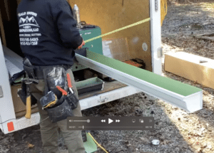 A construction worker in a black hoodie and green pants is seen at the back of a truck, using a machine to cut a long piece of green and white metal. The worker's tool belt is equipped with various tools. The truck, belonging to Halo Roofing, has company details on its door.