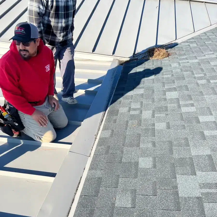 Two workers are on a roof, near the area where a metal roof section meets an asphalt shingle section. One is seated wearing a red hoodie and a cap, and the other is standing in a gray plaid shirt and cap. They appear to be inspecting or discussing the roofing work.