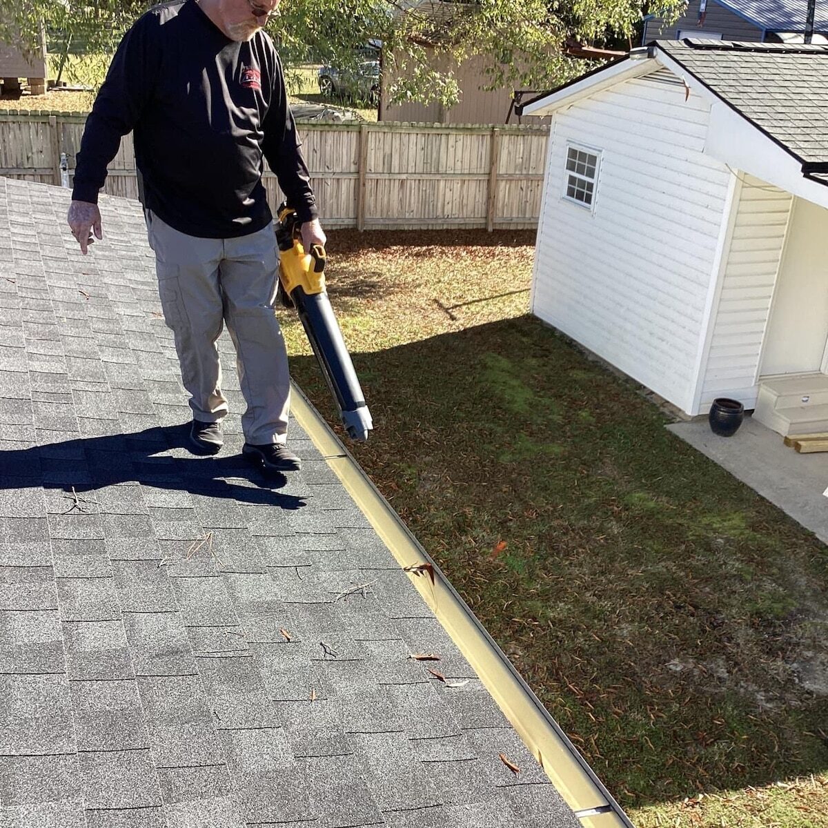 A man, dressed in sunglasses, a black long-sleeve shirt, and gray pants, is using a blower to clear leaves from the gutter of a rooftop. Halo Roofing's meticulous attention to detail ensures every surface is pristine. In the background, a white shed and a fenced backyard with trees are visible.